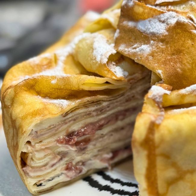 Close-up of a sliced creamy pancake cake showing delicate crepe layers filled with cream cheese, whipped cream, and raspberry jam, wrapped in golden pancakes and dusted with powdered sugar.