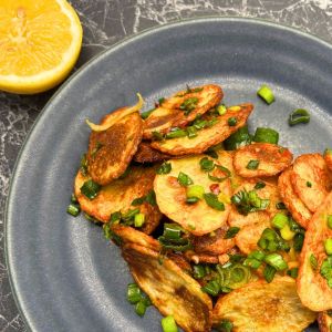 Golden crispy potato slices garnished with chopped green onion on a dark plate, with a halved lemon on the side.