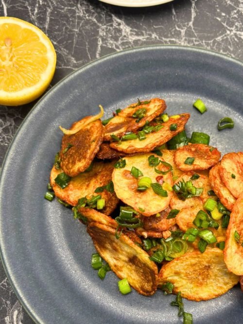 Golden crispy potato slices garnished with chopped green onion on a dark plate, with a halved lemon on the side.