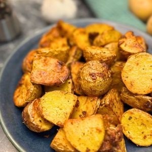 Crispy air fryer roasted potatoes on a dark blue plate, seasoned with herbs and spices, with golden-brown edges and a garlic bulb in the blurred background.