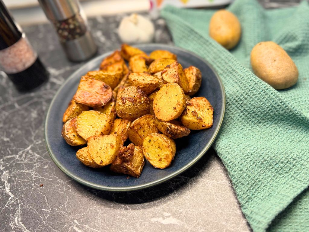 Crispy air fryer potatoes served on a dark plate, placed on a marble counter with whole potatoes, garlic, and a green kitchen towel in the background.