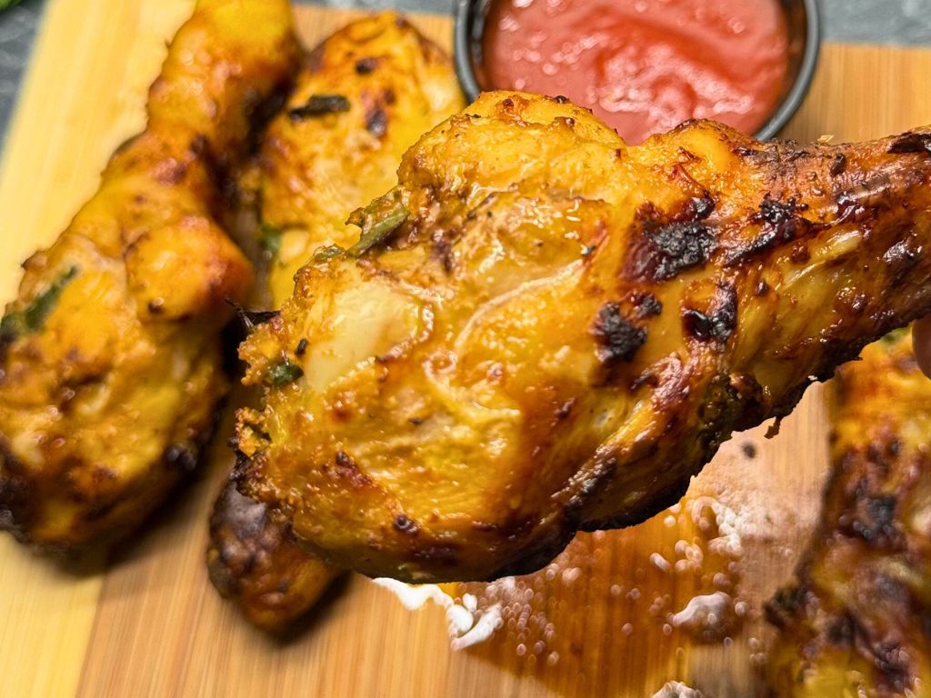 Close-up of a golden, crispy air fryer chicken drumstick held in focus, with more drumsticks and red dipping sauce blurred in the background