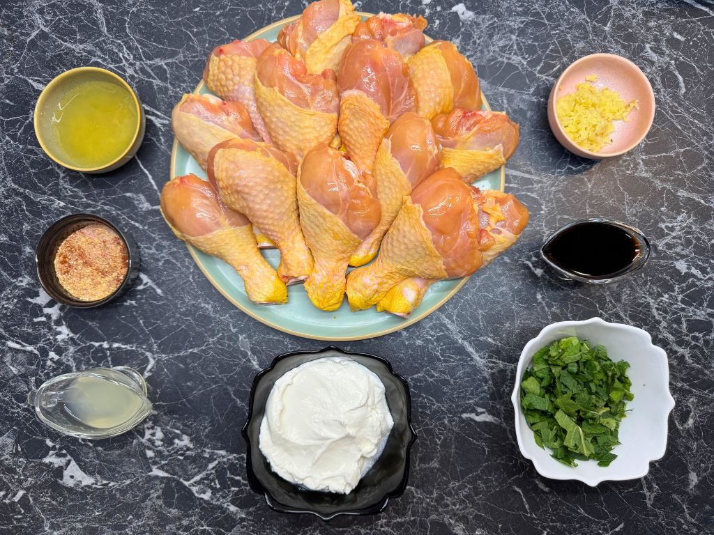 Flat lay of raw chicken drumsticks surrounded by ingredients for a mint marinade, including chopped mint, sour cream, soy sauce, lemon juice, garlic, spices, and honey on a black marble surface.