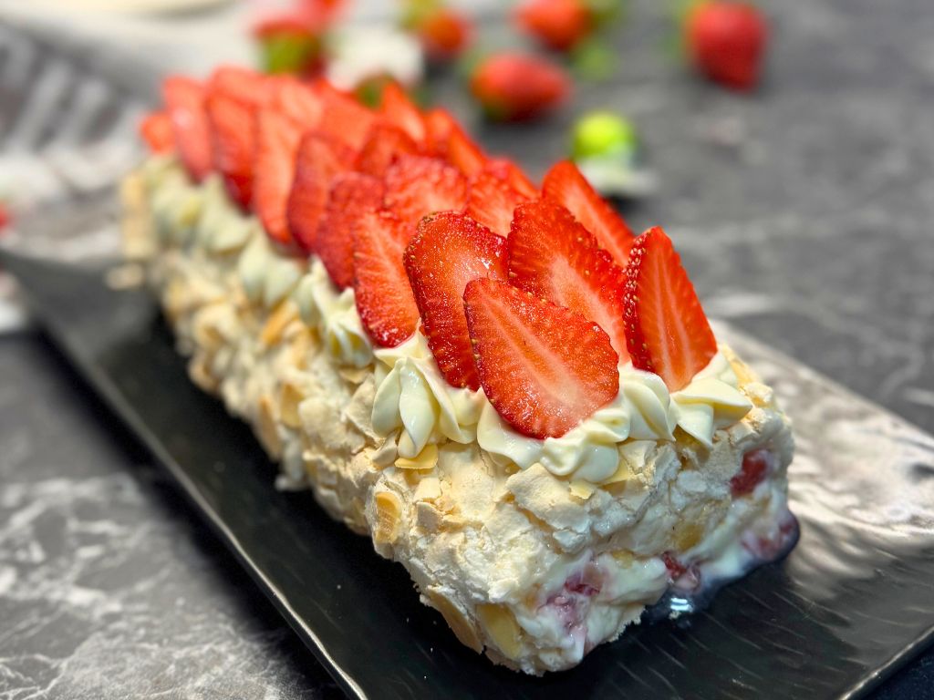 Front angle view of a strawberry meringue roll topped with fresh sliced strawberries and cream cheese swirls, displayed on a black serving platter with scattered strawberries in the background.
