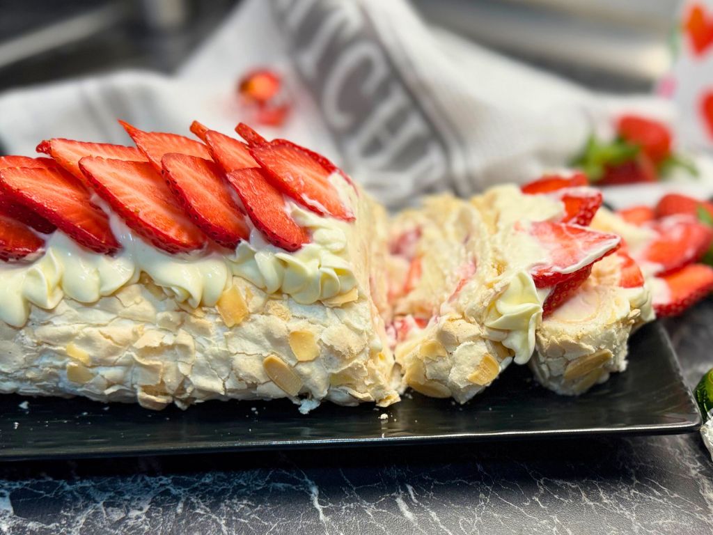 Side view of a sliced strawberry meringue roulade filled with cream and strawberries, topped with piped cream and fresh strawberry slices, served on a black platter with a kitchen towel in the background.