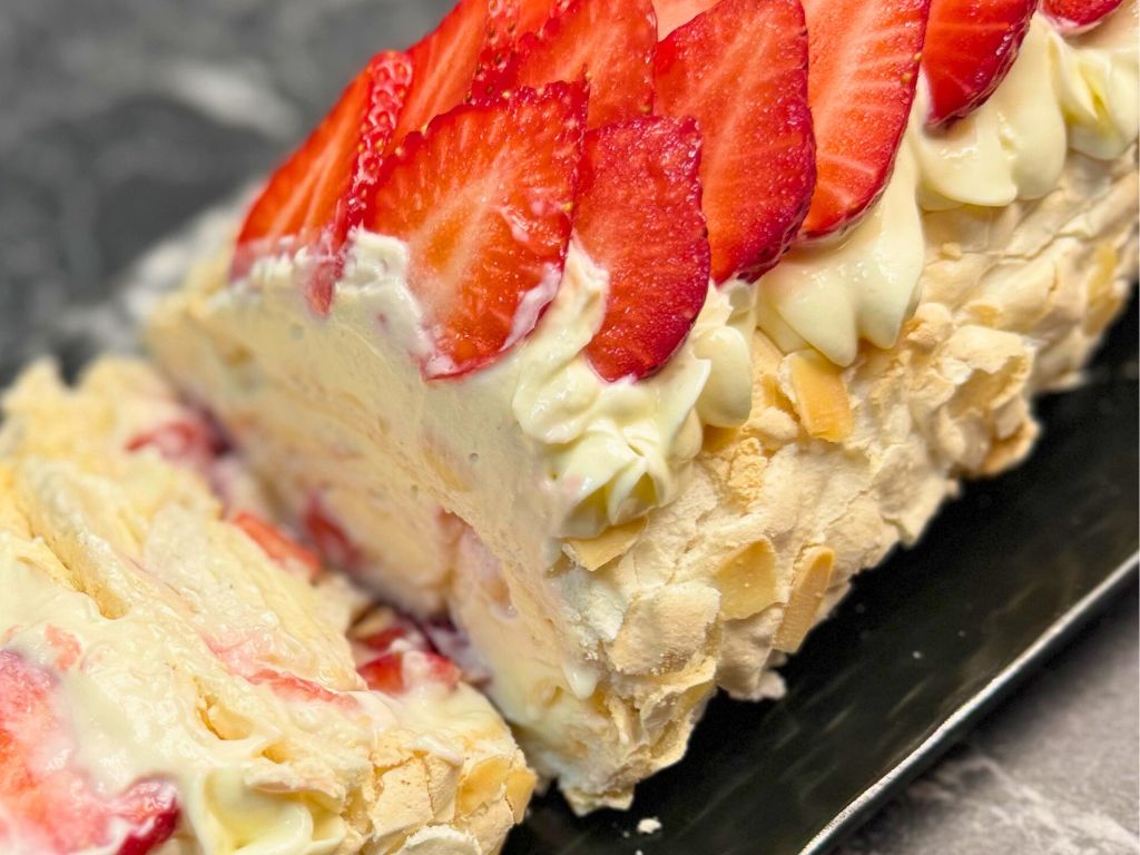 Close-up of a sliced strawberry meringue roll with layers of whipped cream and strawberries, topped with piped cream cheese frosting and fresh strawberry slices on a golden almond crust.