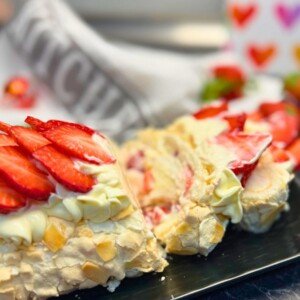 Close-up of a sliced strawberry meringue roll topped with fresh strawberry slices and cream cheese frosting, revealing a light, fluffy meringue filled with whipped cream and strawberries, served on a black platter.