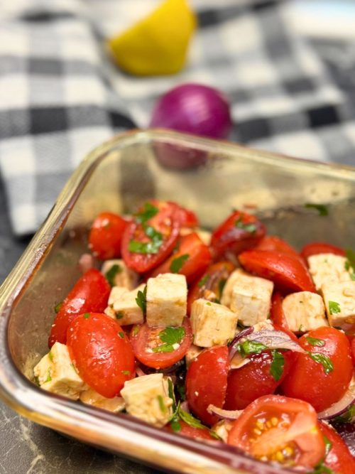 A fresh tomato feta salad in a glass container featuring halved cherry tomatoes, cubed feta cheese, thinly sliced red onions, and chopped parsley, all lightly dressed. In the background, a checkered cloth, a halved lemon, and a whole red onion are slightly out of focus.