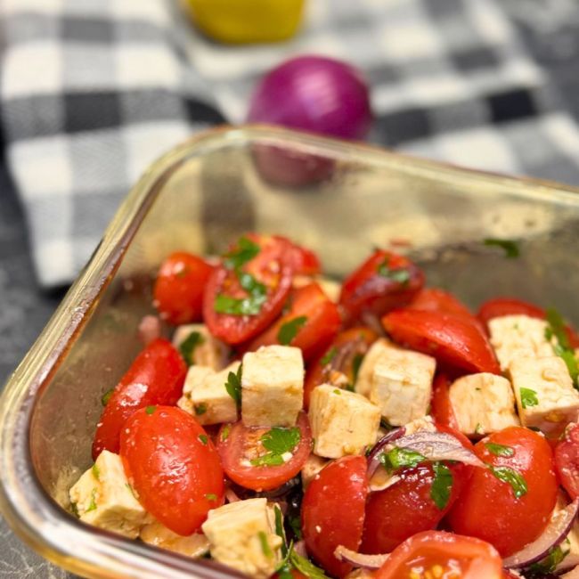 A fresh tomato feta salad in a glass container featuring halved cherry tomatoes, cubed feta cheese, thinly sliced red onions, and chopped parsley, all lightly dressed. In the background, a checkered cloth, a halved lemon, and a whole red onion are slightly out of focus.
