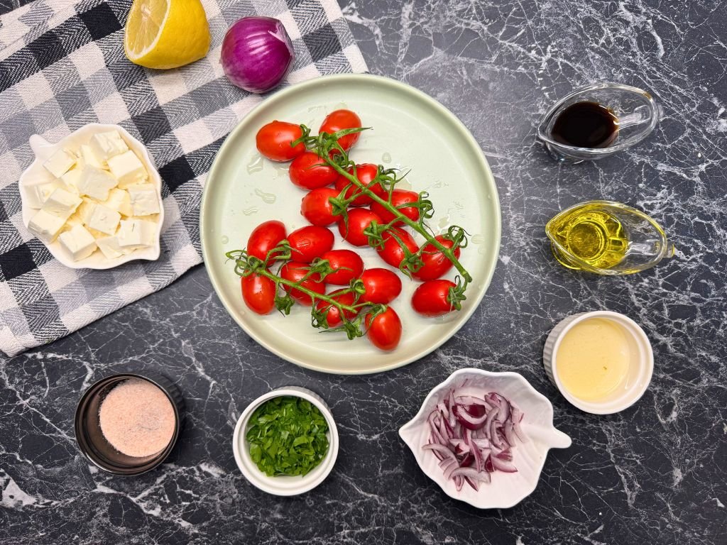 Flat lay of tomato feta salad ingredients on a dark marble countertop, including cherry tomatoes on the vine, feta cubes, red onion, parsley, pink salt, olive oil, soy sauce, and honey.