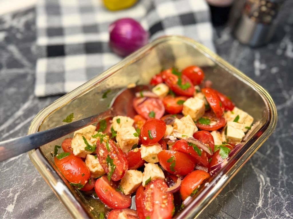 Finished tomato feta salad in a glass container, featuring cherry tomatoes, cubed feta, red onion, and parsley, with a spoon ready for serving