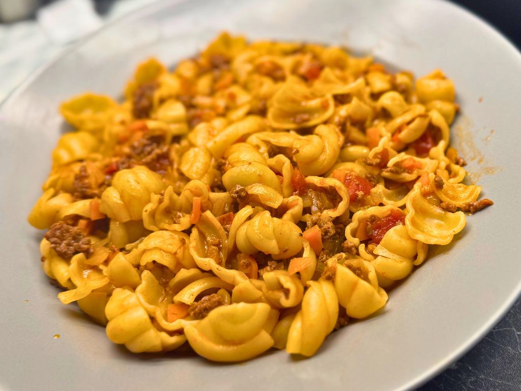 A plate of pasta gnocchi served with Tuscan beef ragu, showing ridged pasta coated in a rich tomato and ground beef sauce with visible chunks of carrot and tomato.