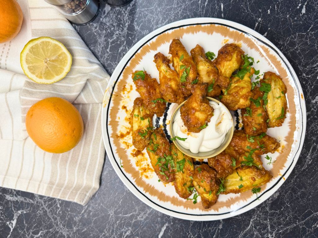 Overhead shot of air fryer orange chicken wings served on a round plate with fresh parsley and a bowl of dipping sauce, surrounded by lemon and orange on a striped towel.