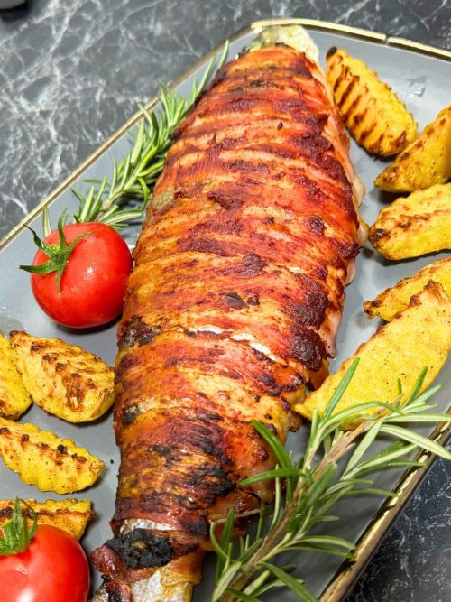 Close-up of bacon-wrapped trout served with golden potato wedges, fresh cherry tomatoes, and rosemary sprigs on a gray plate