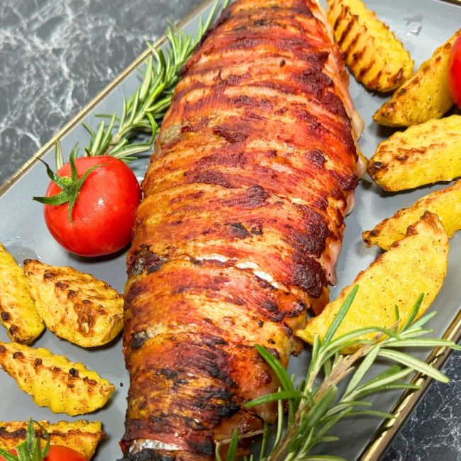 Close-up of bacon-wrapped trout served with golden potato wedges, fresh cherry tomatoes, and rosemary sprigs on a gray plate