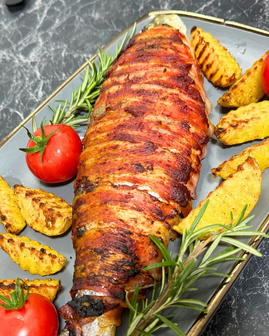 Close-up of bacon-wrapped trout served with golden potato wedges, fresh cherry tomatoes, and rosemary sprigs on a gray plate