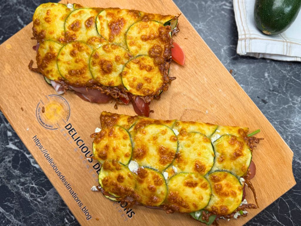 Two baked zucchini mozzarella sandwiches on a wooden board, filled with spinach, tomato, cream cheese, and prosciutto, ready to serve.