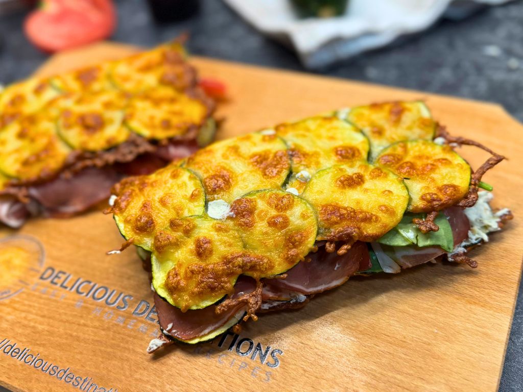 Cheesy baked zucchini sandwich on a wooden board, filled with spinach, tomato, cream cheese, and prosciutto, with golden melted mozzarella on top.