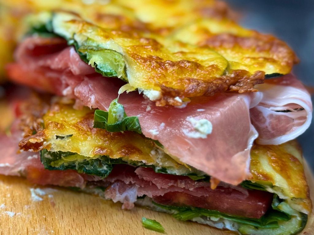 Stacked slices of baked zucchini sandwich with crispy mozzarella top, layered with fresh spinach, tomato, cream cheese, and prosciutto.