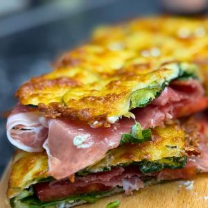 Close-up of a baked zucchini sandwich with golden mozzarella crust, layered with cream cheese, spinach, tomatoes, and prosciutto on a wooden board.