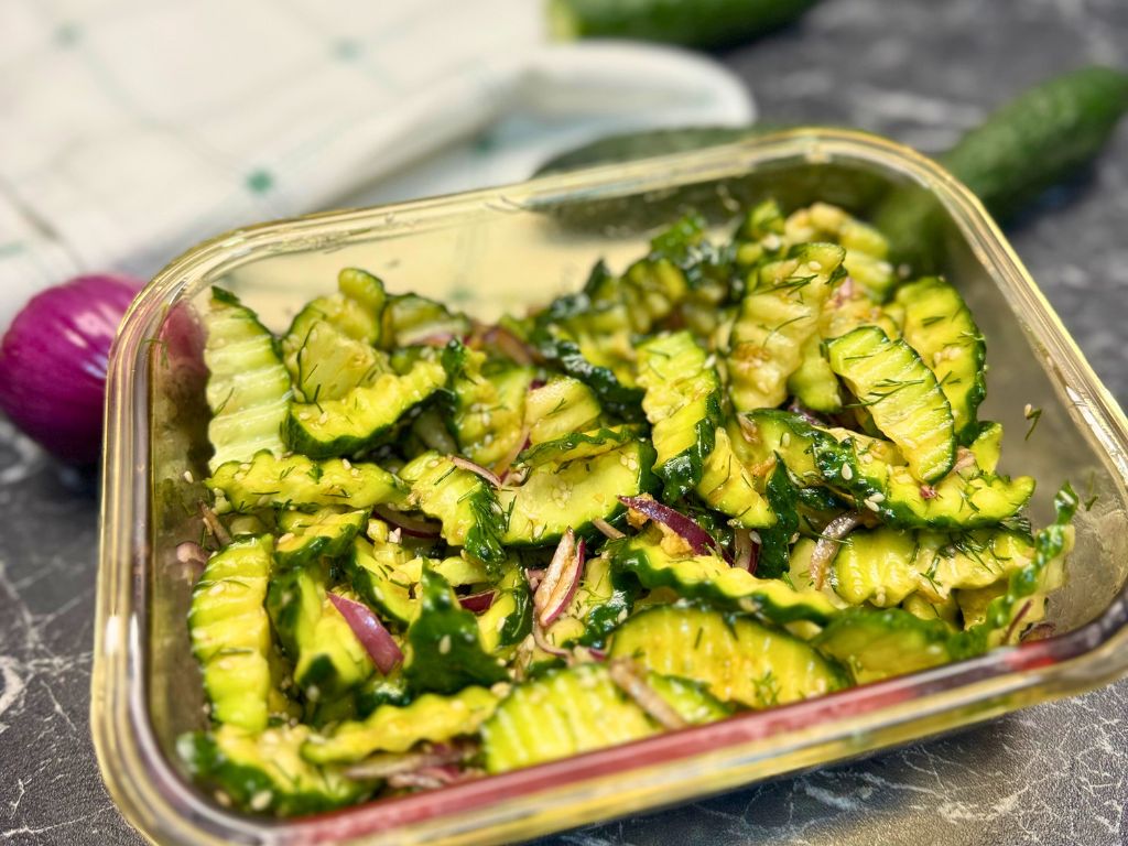 Glass container filled with garlic cucumber salad featuring crinkle-cut cucumbers, red onion, dill, and sesame seeds on a dark countertop.