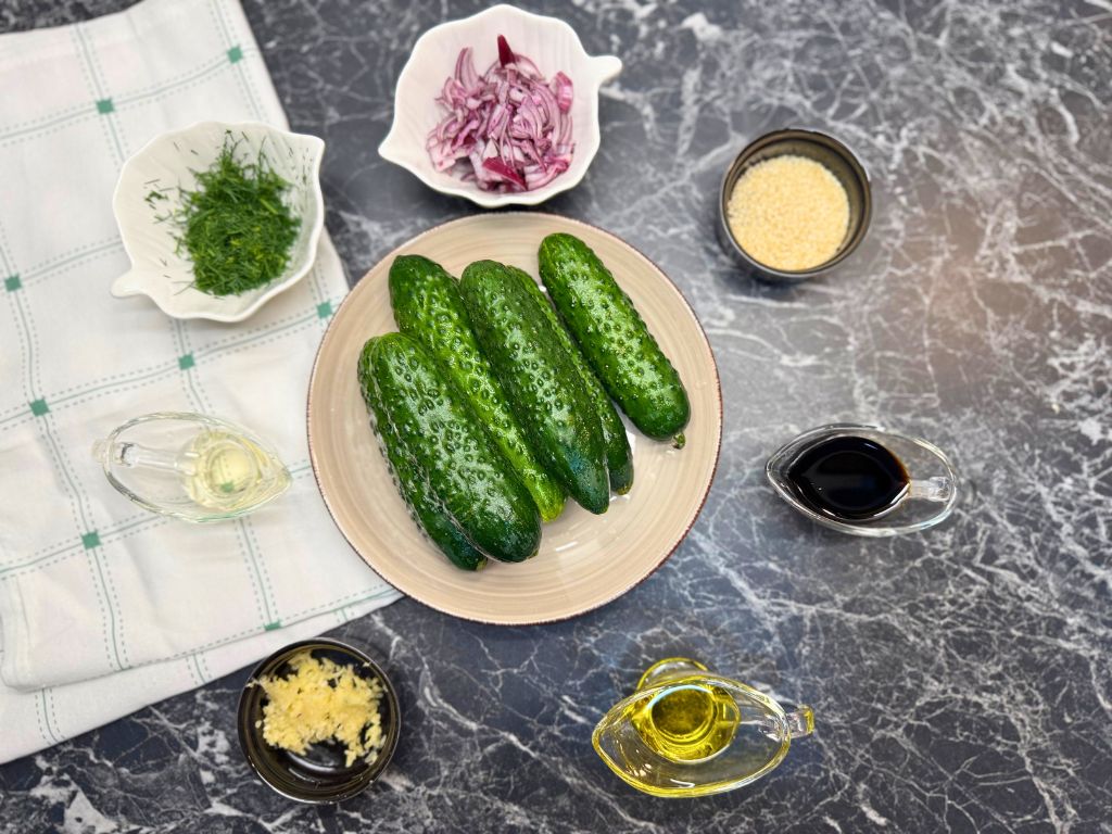 Fresh ingredients for garlic cucumber salad recipe, including cucumbers, red onion, dill, sesame seeds, soy sauce, vinegar, oil, and garlic.