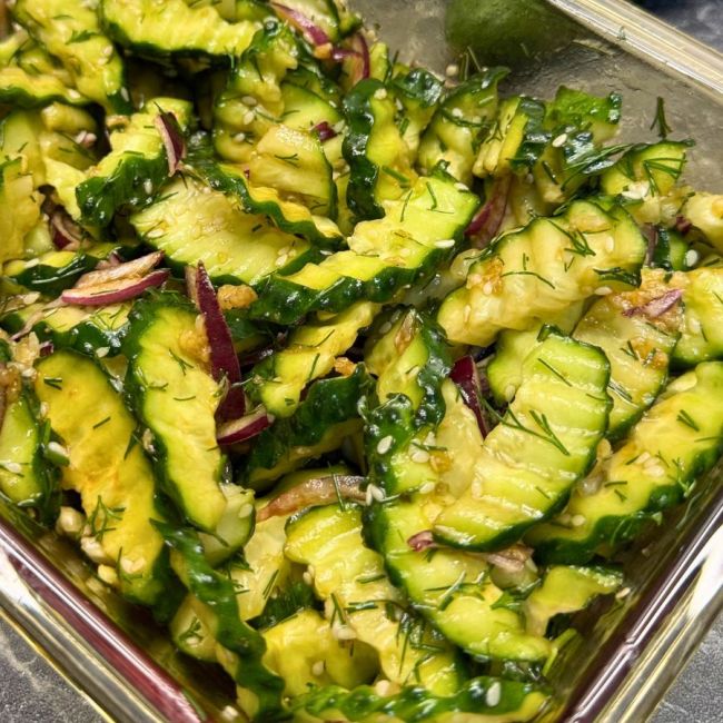 Garlic cucumber salad with red onion, dill, sesame seeds, and soy dressing in a glass container.