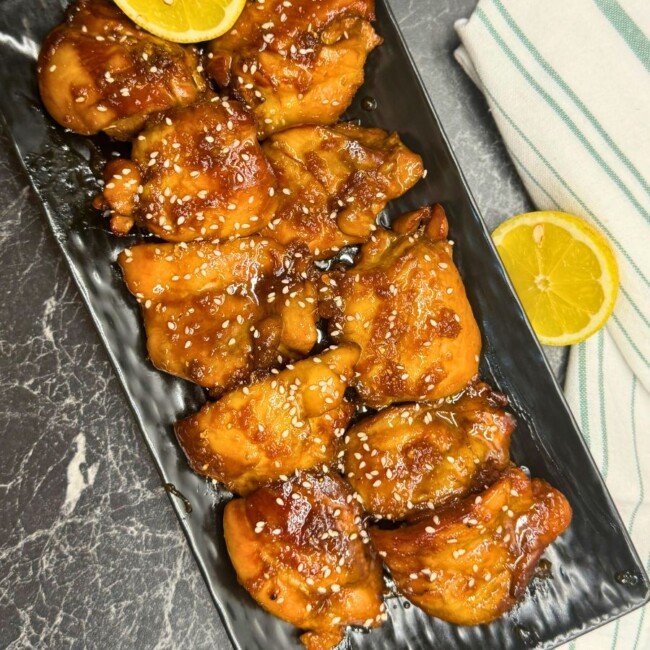 Glazed Lemon Teriyaki Chicken Thighs topped with sesame seeds and lemon slices on a black serving platter.