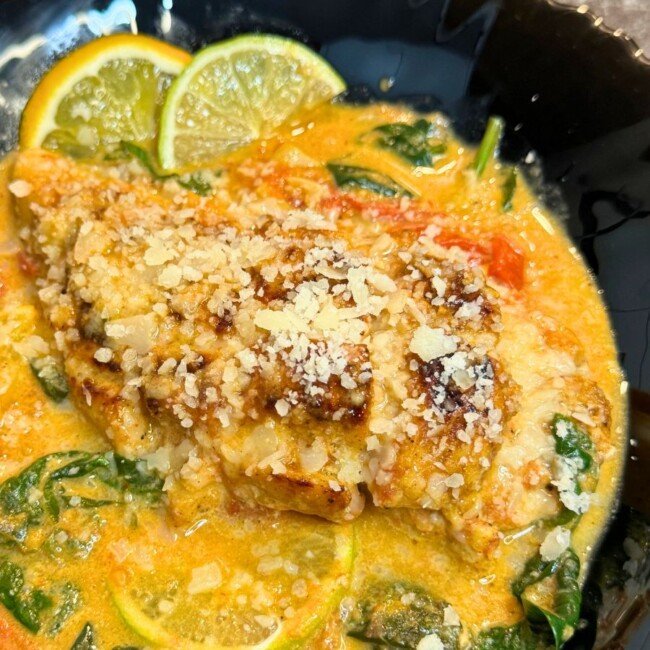 Pan-seared chicken breast topped with grated cheese, served in a creamy orange-colored sauce with spinach, cherry tomatoes, and lemon slices, presented in a black bowl.