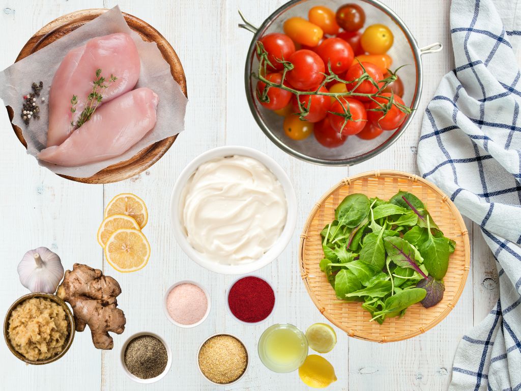 Flat lay of ingredients for creamy chicken: raw chicken breasts, cherry tomatoes, fresh spinach, sour cream, lemon slices, garlic, ginger, and small bowls of spices on a white wooden surface with a kitchen towel.