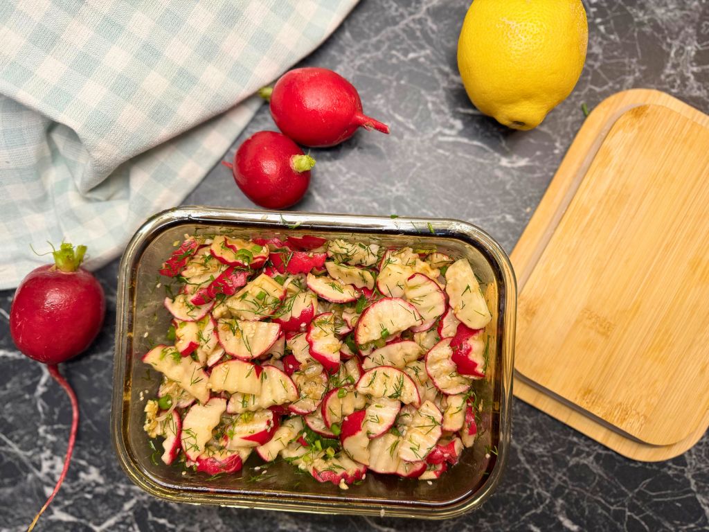 Overhead view of marinated pickled radish with dill in a glass container, surrounded by fresh radishes, a lemon, and a wooden cutting board on a dark countertop.