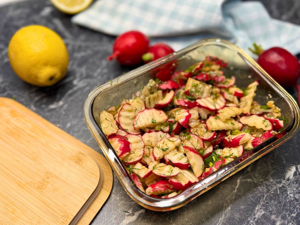 Angled view of quick pickled radish in a glass container, garnished with dill and green onion, with fresh radishes and lemon in the background on a dark kitchen counter.
