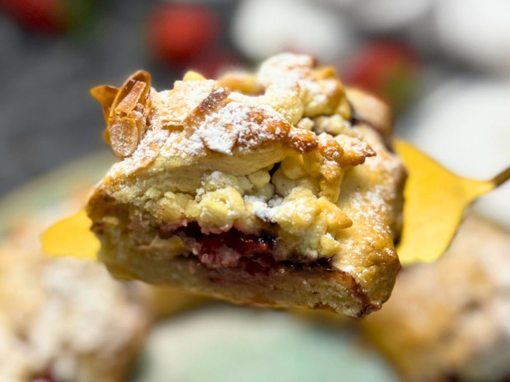 Close-up of a slice of strawberry tart on a gold dessert fork, showing the crumbly crust, juicy strawberry filling, toasted almonds, and powdered sugar topping.
