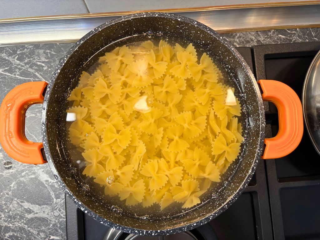 A pot of bowtie pasta (farfalle) boiling in water on the stove, with orange handles and visible steam starting to rise.