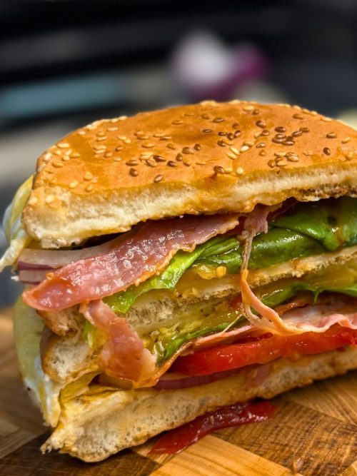 Close-up of a cheesy bacon egg burger cut in half, showing layers of bacon, lettuce, tomato, onion, cheese, and fluffy egg in a sesame bun.