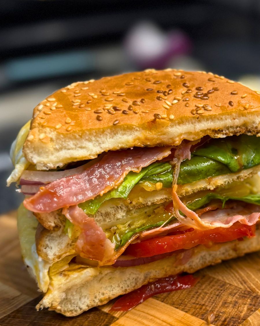 Close-up of a cheesy bacon egg burger cut in half, showing layers of bacon, lettuce, tomato, onion, cheese, and fluffy egg in a sesame bun.