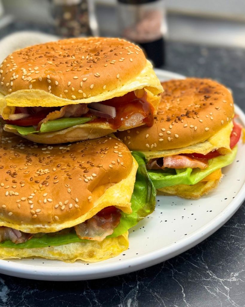 Three cheesy bacon egg burgers served on a white plate, each stacked with lettuce, bacon, egg, and tomato in toasted sesame buns.