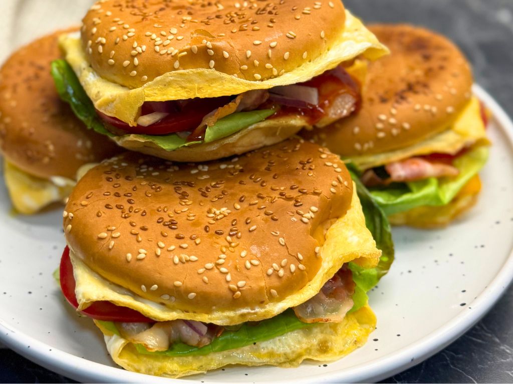 Four cheesy bacon egg burgers on a white plate, each filled with lettuce, bacon, tomato, red onion, and melted cheese wrapped in egg.