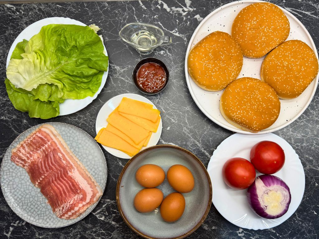 Ingredients for cheesy bacon egg burgers, including lettuce, bacon, eggs, cheddar slices, burger buns, tomato, red onion, sauce, and oil on a kitchen counter.