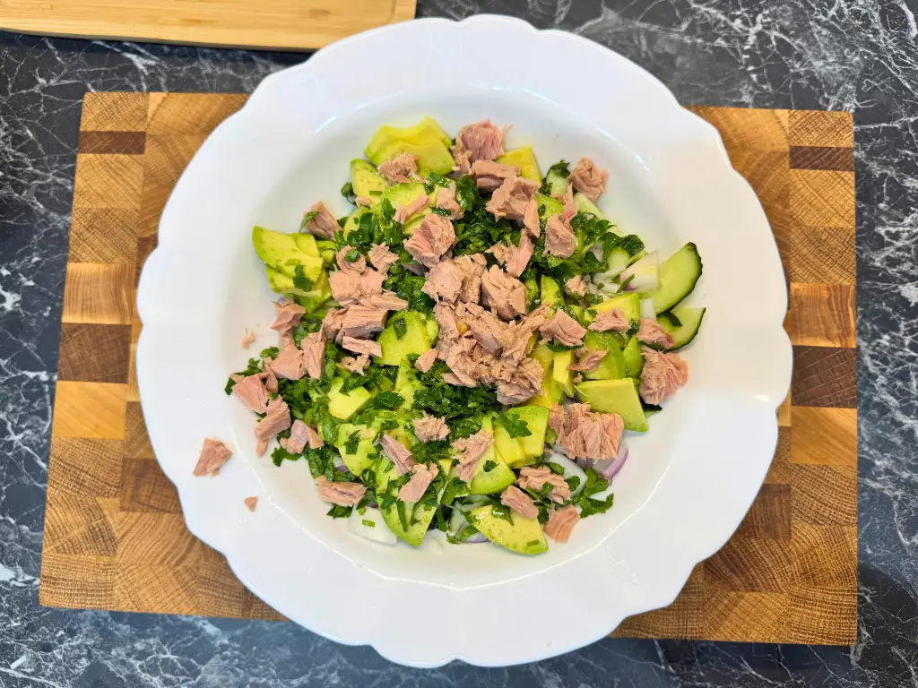 Mixing cucumber, avocado, parsley, and tuna in a white bowl to make a healthy avocado tuna cucumber salad.