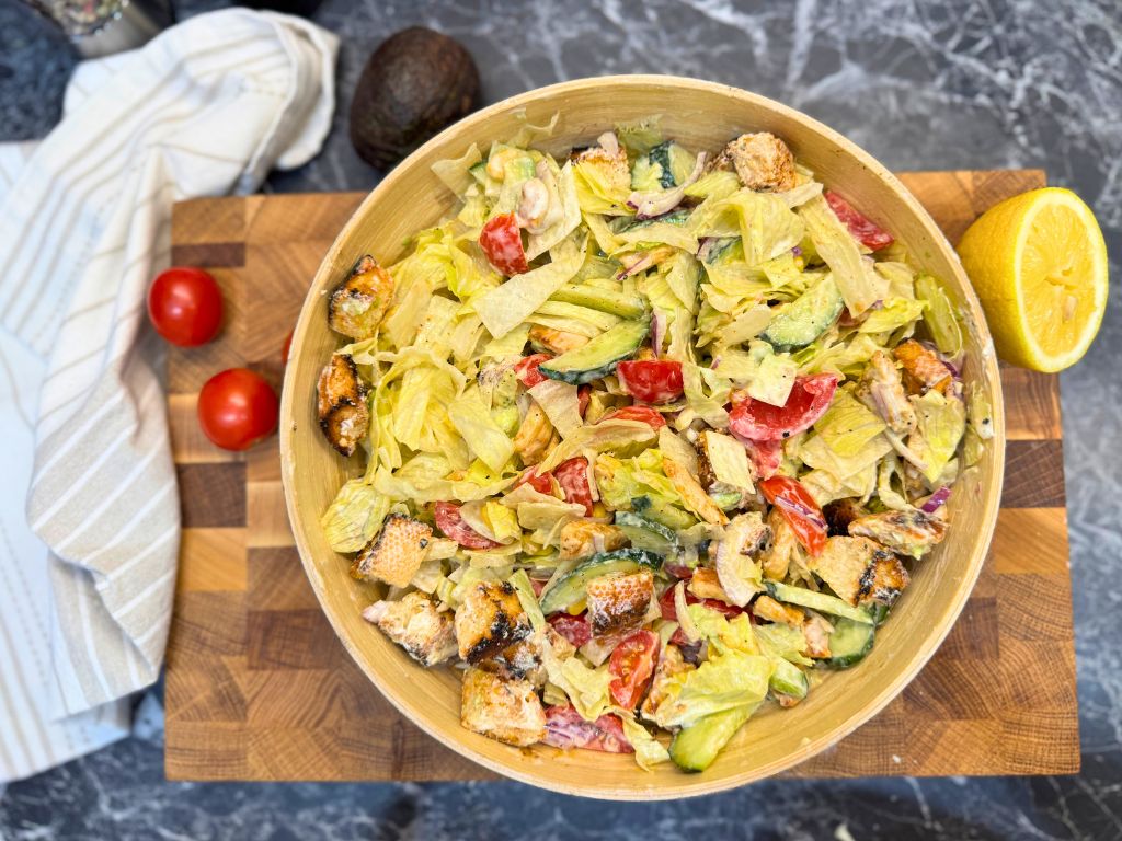 Bowl of healthy chicken salad with avocado, lettuce, cherry tomatoes, cucumber, croutons, and creamy garlic dressing on a wooden board
