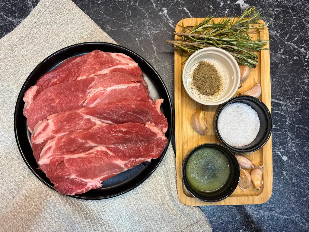 Fresh pork neck slices on a black plate with rosemary sprigs, garlic cloves, salt, black pepper, and melted butter arranged on a wooden tray.