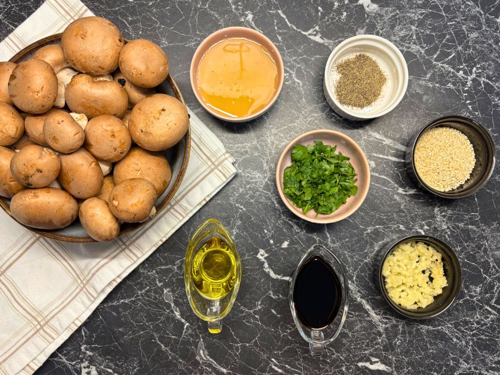 Overhead view of ingredients for soy and honey roasted mushrooms, including fresh cremini mushrooms, garlic, soy sauce, olive oil, honey, parsley, sesame seeds, and black pepper, arranged on a dark marble surface.