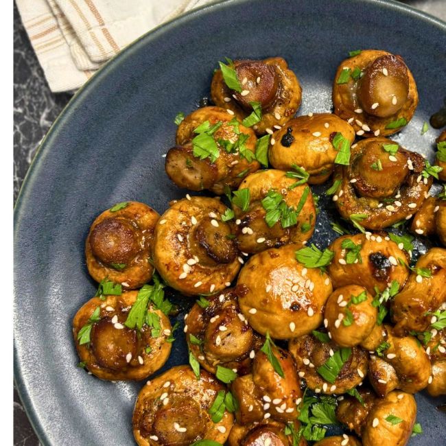 Roasted mushrooms glazed with soy sauce and honey, garnished with sesame seeds and fresh parsley on a dark plate.