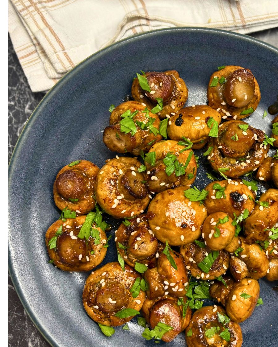 Roasted mushrooms glazed with soy sauce and honey, garnished with sesame seeds and fresh parsley on a dark plate.