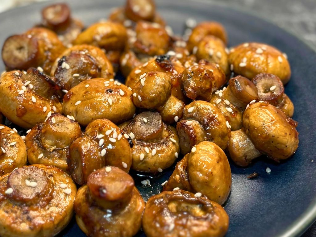 Close-up of caramelized roasted mushrooms glazed with soy sauce and honey, sprinkled with sesame seeds on a dark plate.