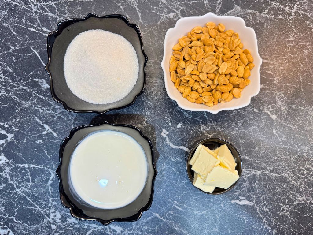 Ingredients for caramel peanut topping including granulated sugar, roasted peanuts, butter, and heavy cream on a marble surface.