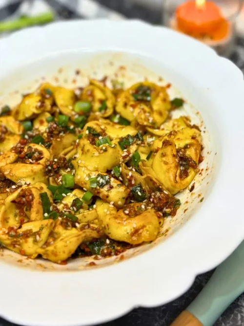 Side view of crispy tortelloni coated in spicy chili oil, sesame, and scallions served in a white bowl.