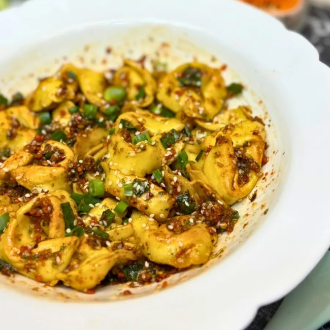 Side view of crispy tortelloni coated in spicy chili oil, sesame, and scallions served in a white bowl.