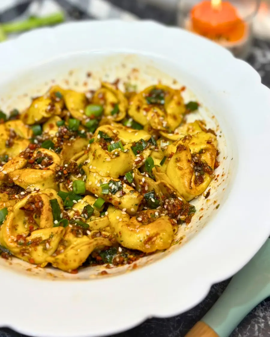 Side view of crispy tortelloni coated in spicy chili oil, sesame, and scallions served in a white bowl.